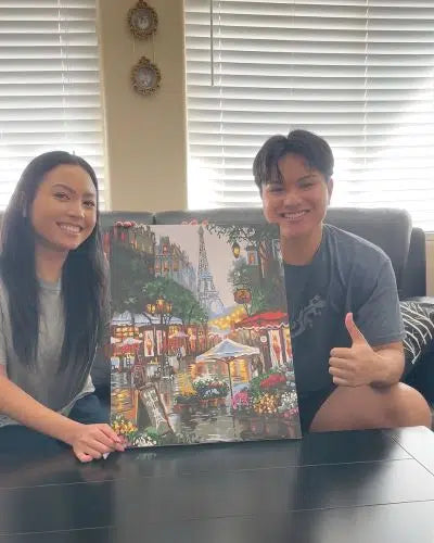 Two people holding a colorful painting of a street scene with buildings and trees.