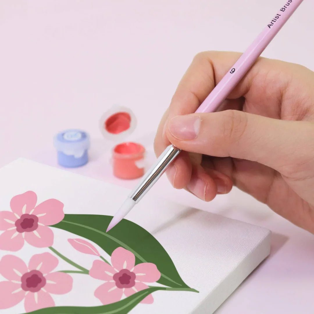 Hand holding a paintbrush near flowers painted on paper with small paint containers.