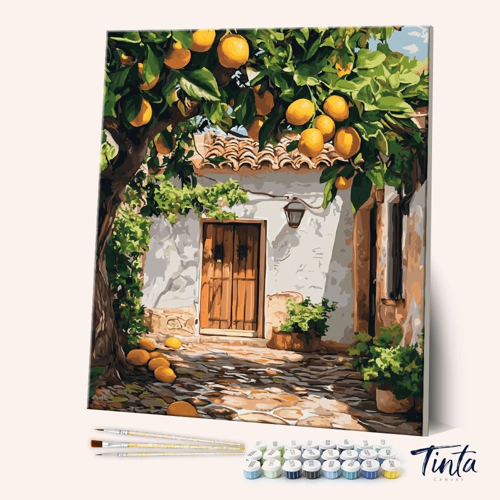 Angled view of Lemon Yard paint by numbers kit showing a sunlit cottage beneath lemon trees with brushes and paints by Tinta Canvas