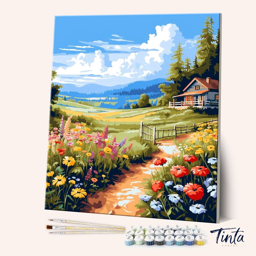 Angled display of the Meadow Cottage paint by numbers kit showing cottage landscape canvas with brushes and paint pots by Tinta Canvas