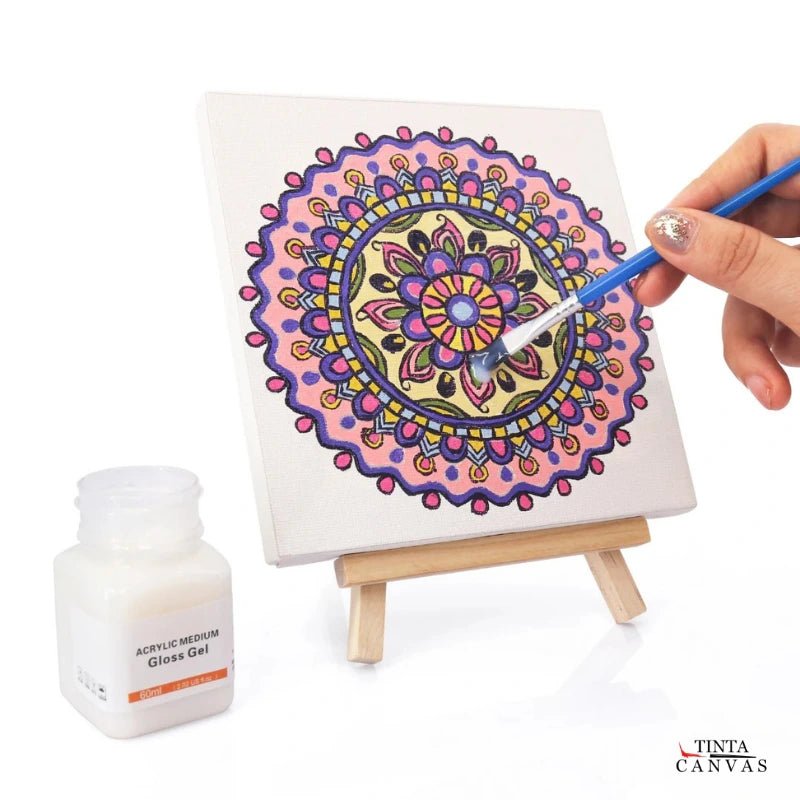 Person painting a colorful mandala on a small canvas with a Primer Gesso bottle of acrylic medium next to it.